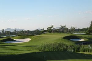Van Tri Golf Club green guarded by deep pot bunkers, with tall grass and distant hills near Hanoi
