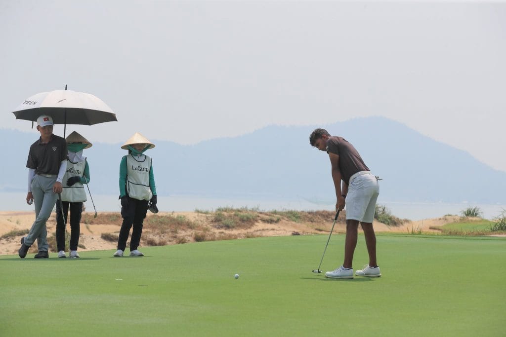 Golfer putting on the green at Laguna Golf Lang Co in Central Vietnam, with caddies in conical hats waiting beside the green