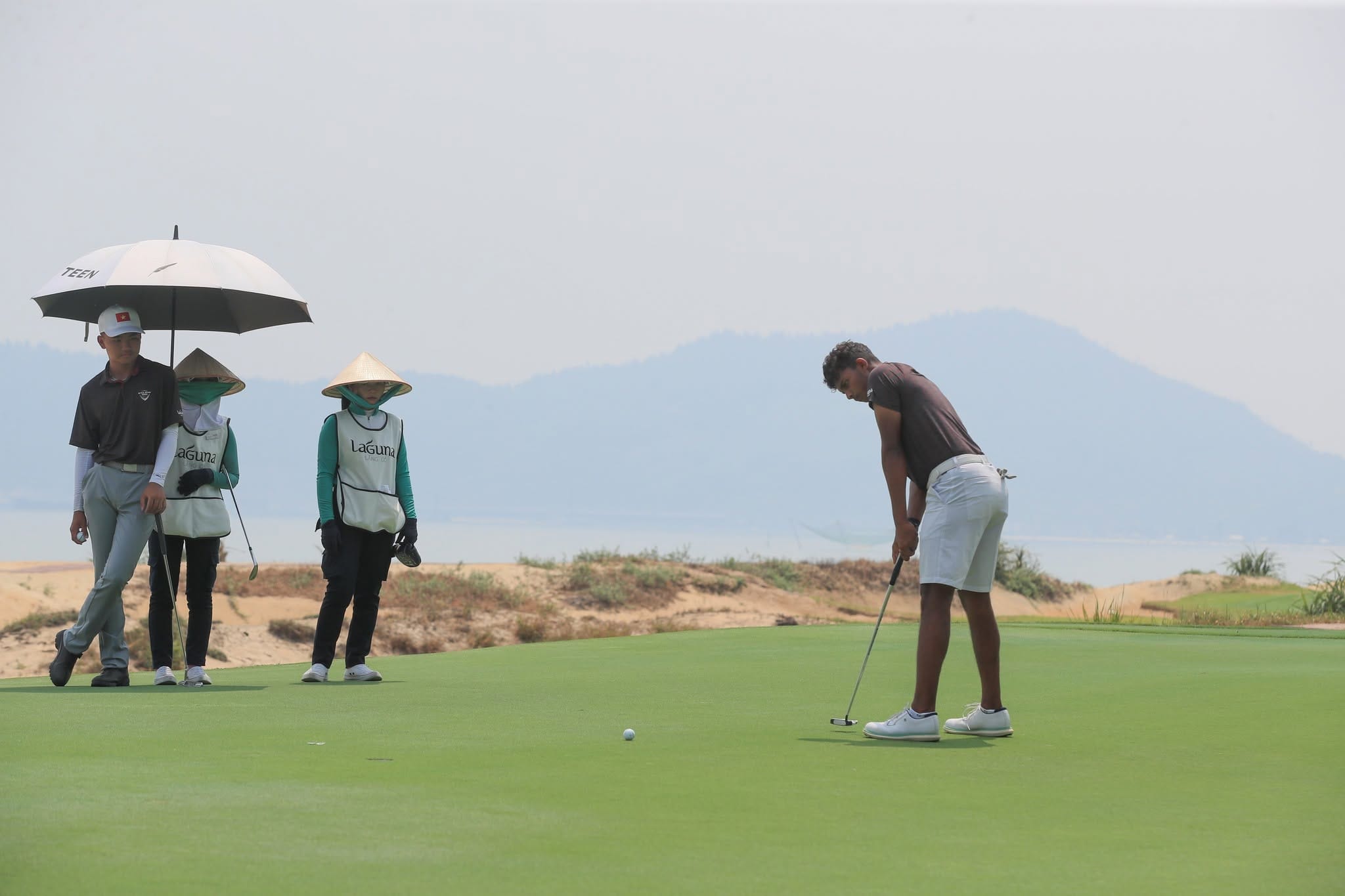 Golfer putting on the green at Laguna Golf Lang Co in Central Vietnam, with caddies in conical hats waiting beside the green