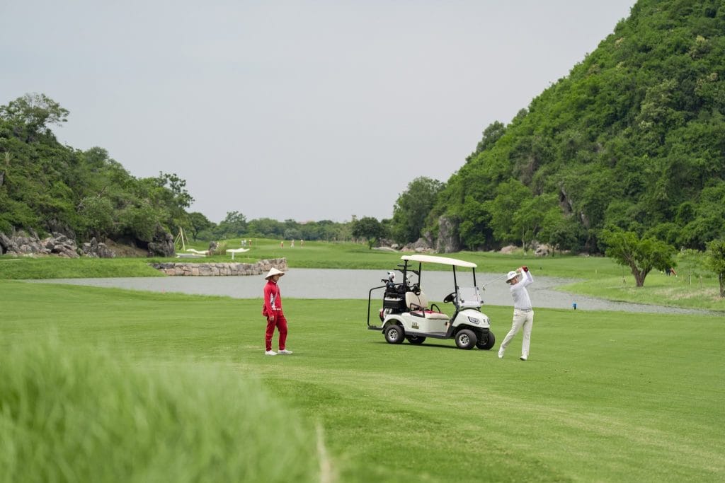 Golfer teeing off with a caddie at BRG golf course near Hanoi, limestone hills and lake behind on a clear spring day