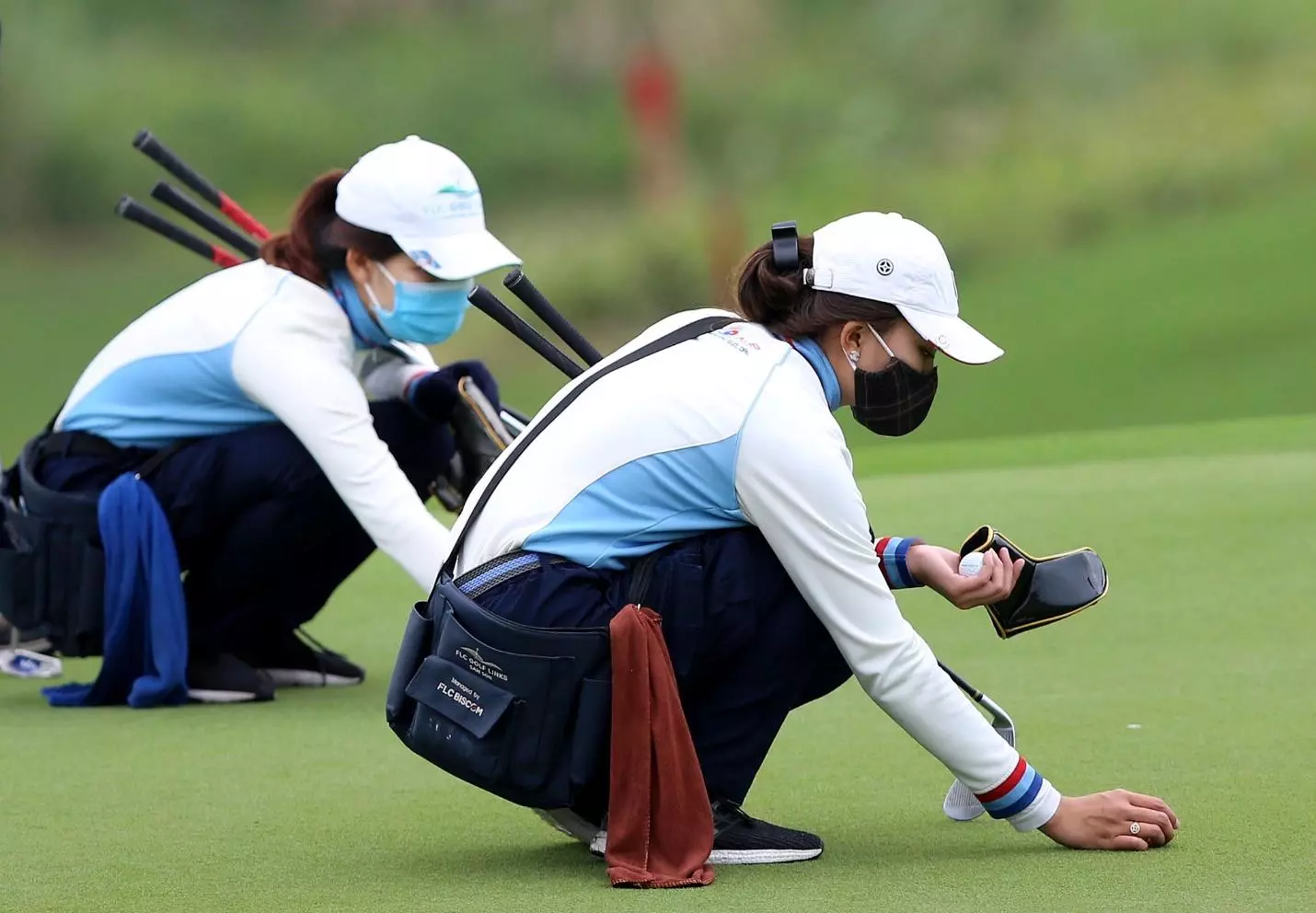 Caddies marking balls on the green Two caddies crouching on a putting green, one placing a ball and holding a putter cover, at a golf course in Vietnam