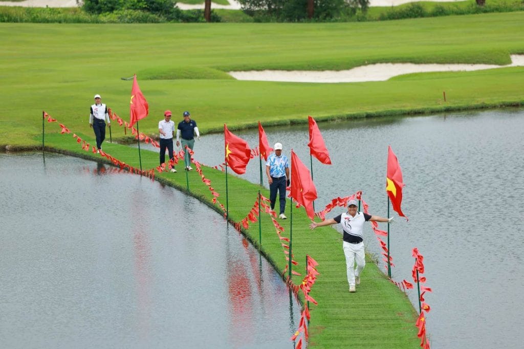 Golfers crossing a lake on a grass walkway lined with Vietnamese flags at a golf course in Vietnam