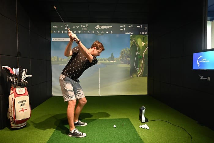 Golfer mid-swing in an indoor golf simulator bay, with a projected course and shot data on the screen