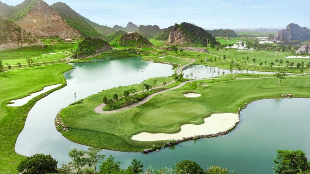 Aerial view of a golf course with greens and bunkers wrapped around a lake, set among limestone karst hills south of Hanoi