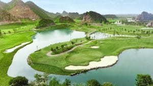 Aerial view of a golf course with greens and bunkers wrapped around a lake, set among limestone karst hills south of Hanoi