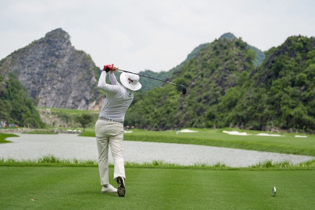 Golfer teeing off across a lake toward limestone hills at a golf course in northern Vietnam