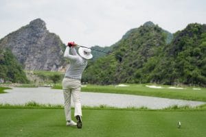 Golfer teeing off across a lake toward limestone hills at a golf course in northern Vietnam