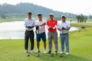Four golfers posing with their drivers on a tee box beside a lake at a golf course near Hanoi